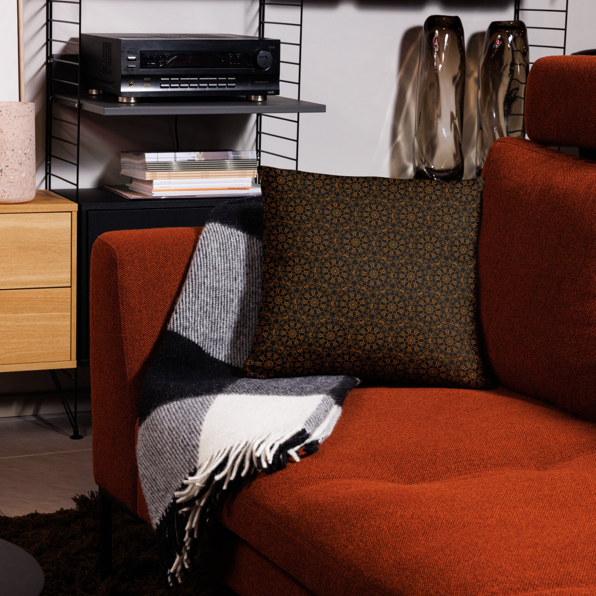 18″×18″ throw pillow with the 'Enneagon' black & gold geometric pattern on a red armchair with checkered blanket.