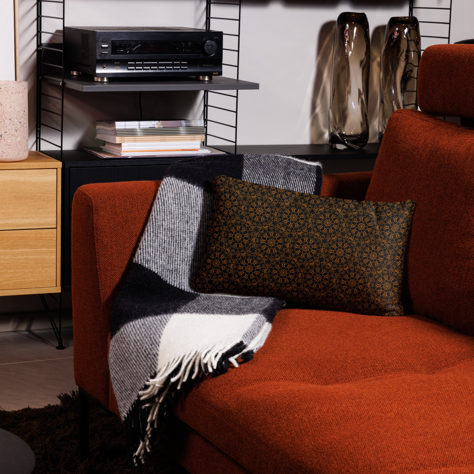 20″×12″ throw pillow with the 'Enneagon' black & gold geometric pattern on a red armchair with checkered blanket.