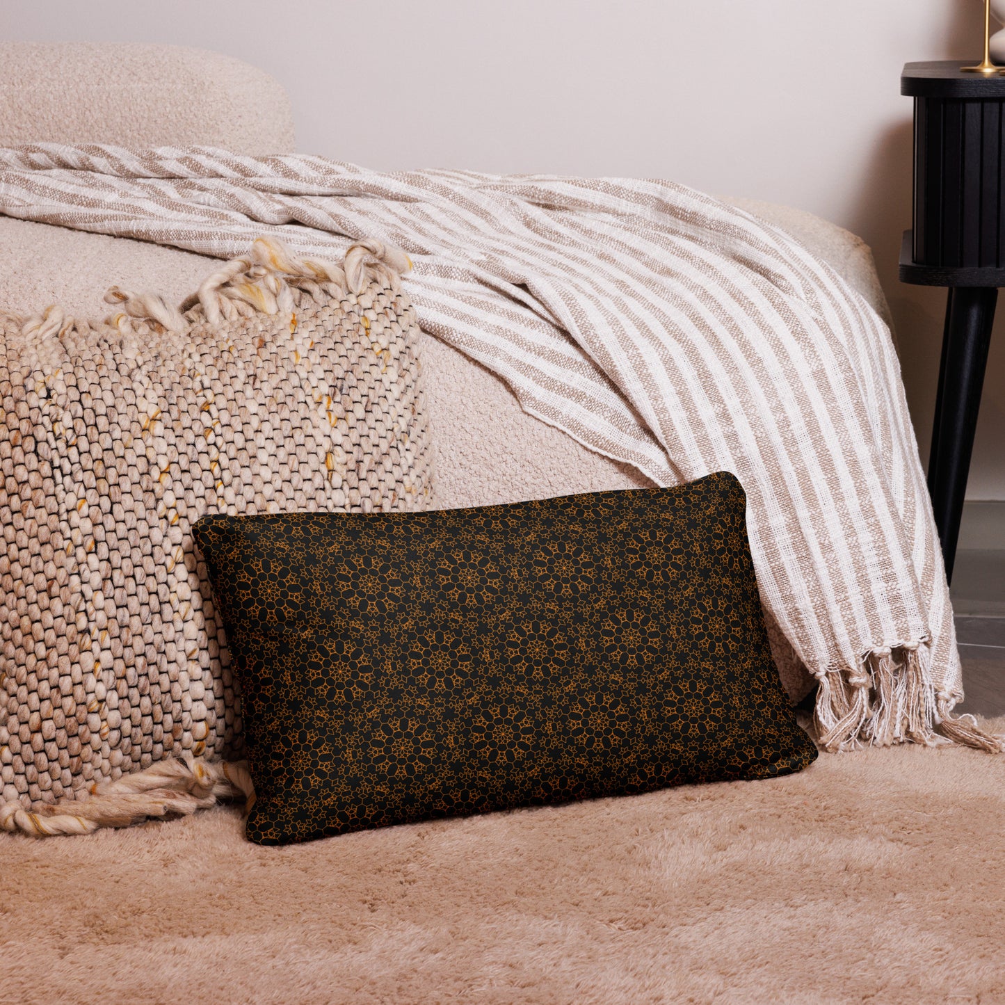 20″×12″ throw pillow with the 'Enneagon' black & gold geometric pattern on a bed with striped blanket.