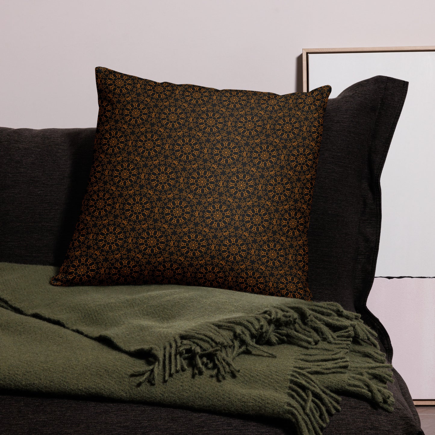 22″×22″ throw pillow with the 'Enneagon' black & gold geometric pattern on a dark sofa with green throw blanket.