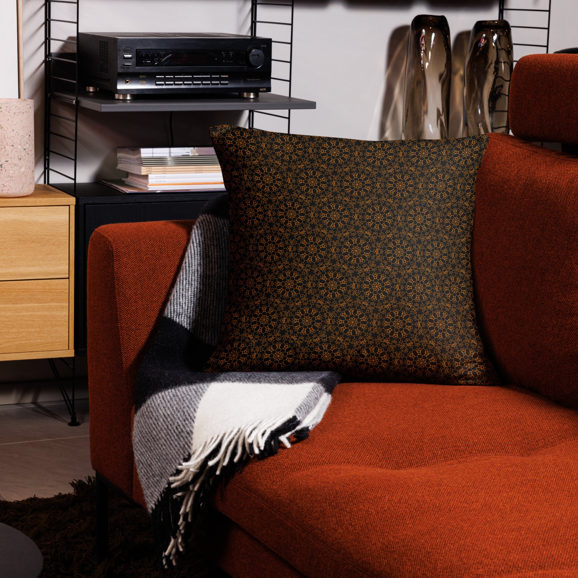 22″×22″ throw pillow with the 'Enneagon' black & gold geometric pattern on a red armchair with checkered blanket.
