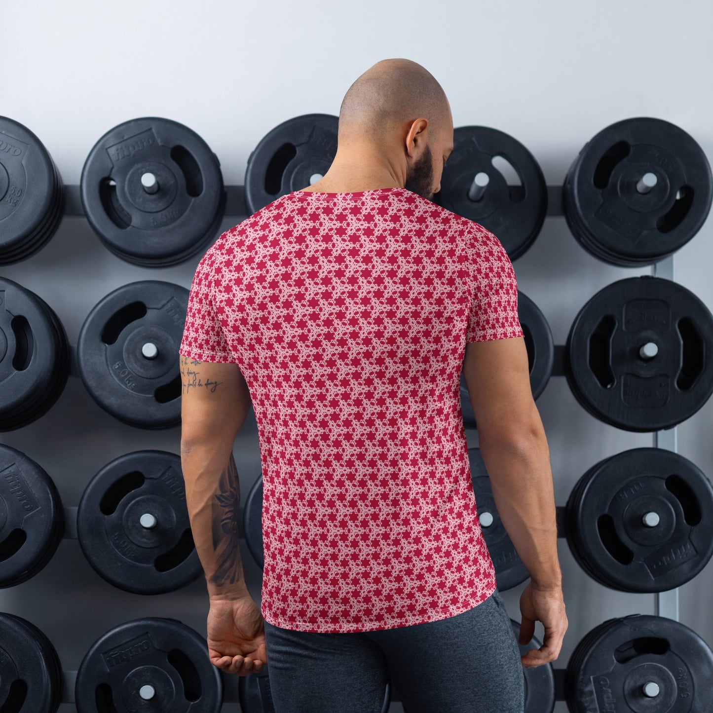 Back shot of an athletic man standing in front of a gym weight plate rack wearing the t-shirt with the 'Trigon' red and pink fractal geometric pattern.
