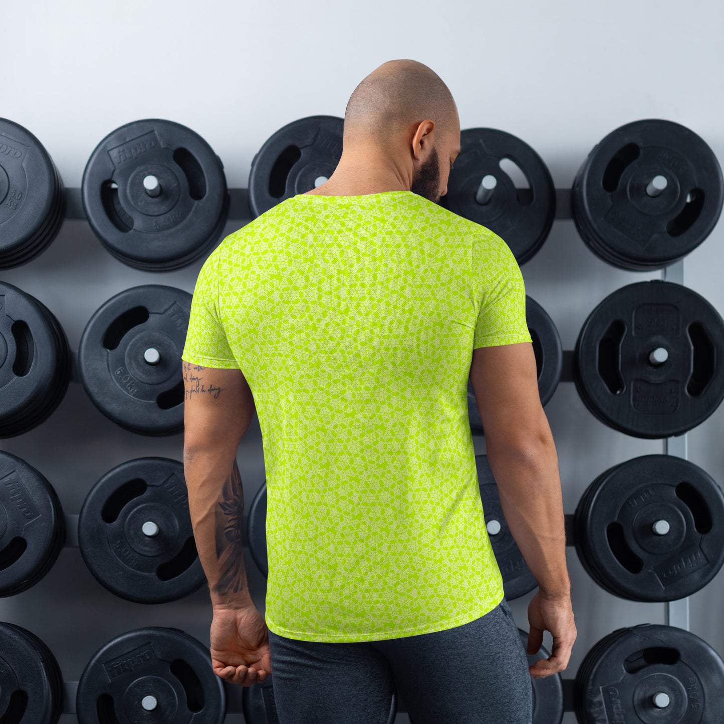 Back shot of an athletic man standing in front of a gym weight plate rack wearing the t-shirt with the 'Heptagon' yellow-green fractal geometric pattern.