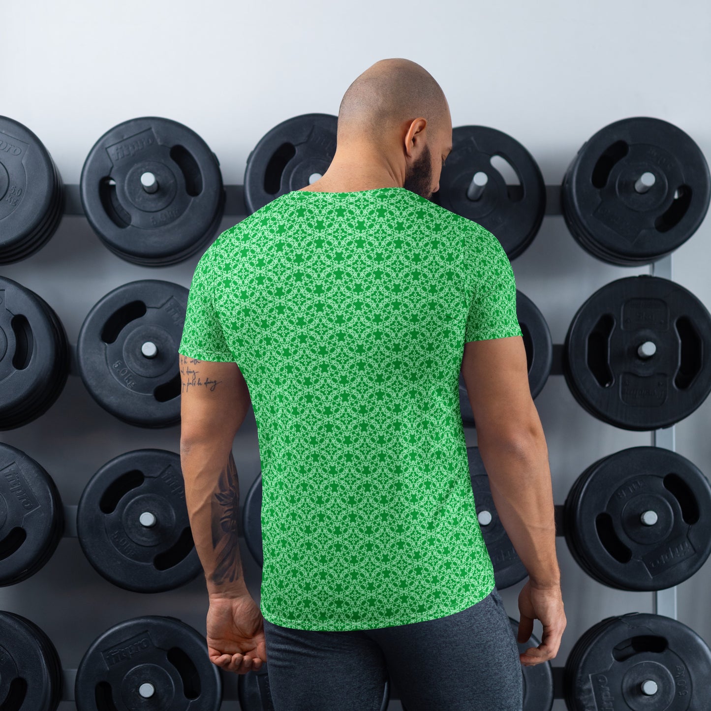 Back shot of an athletic man standing in front of a gym weight plate rack wearing the t-shirt with the 'Tetragon' green fractal geometric pattern.