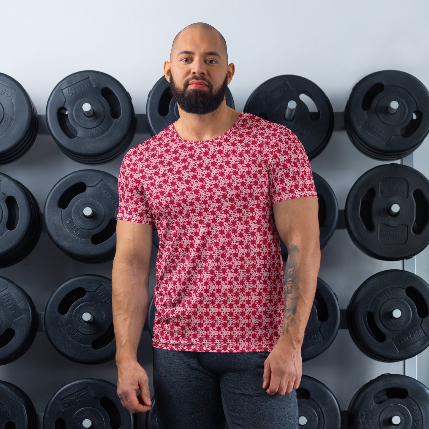Athletic man standing in front of a gym weight plate rack wearing the t-shirt with the 'Trigon' red and pink fractal geometric pattern.