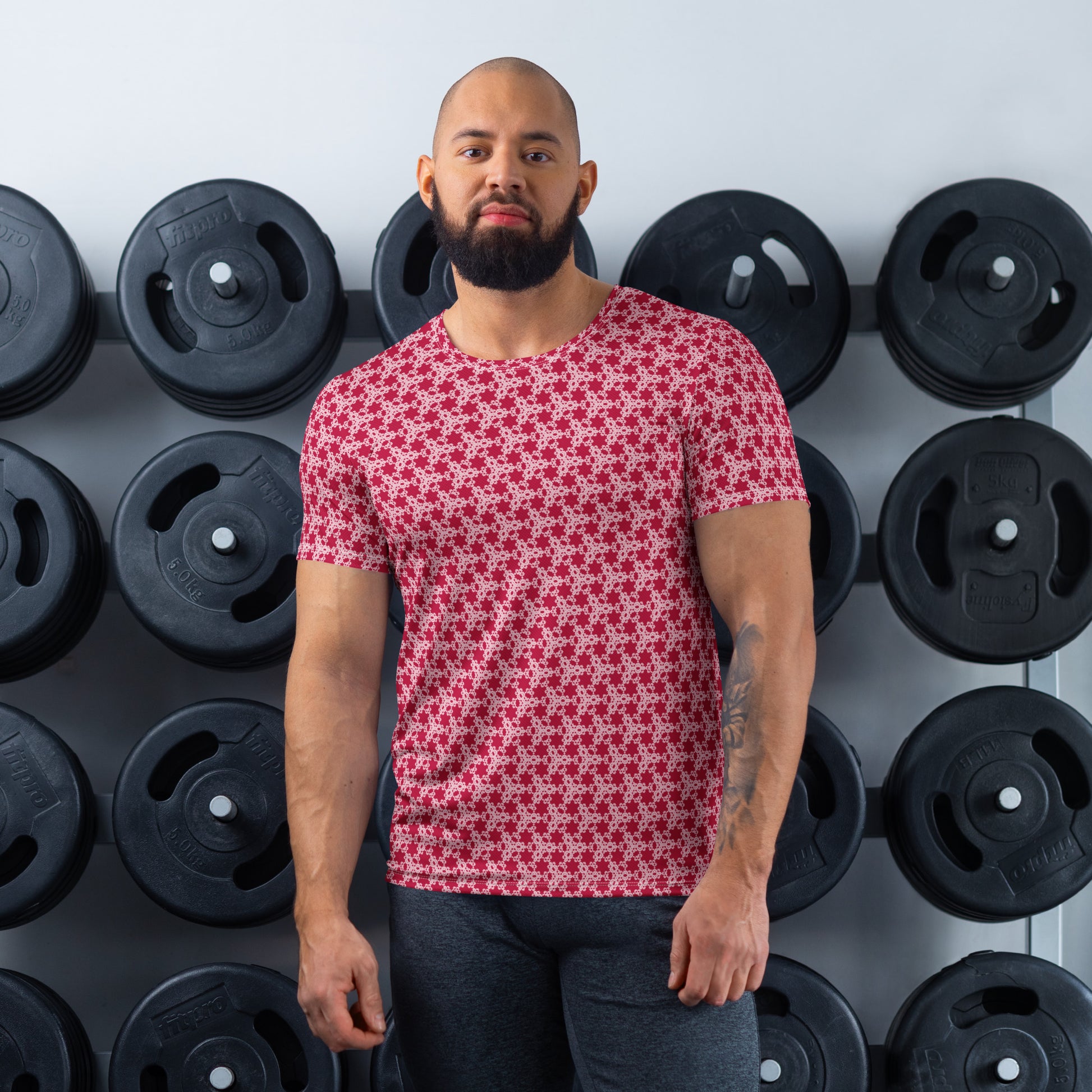 Athletic man standing in front of a gym weight plate rack wearing the t-shirt with the 'Trigon' red and pink fractal geometric pattern.