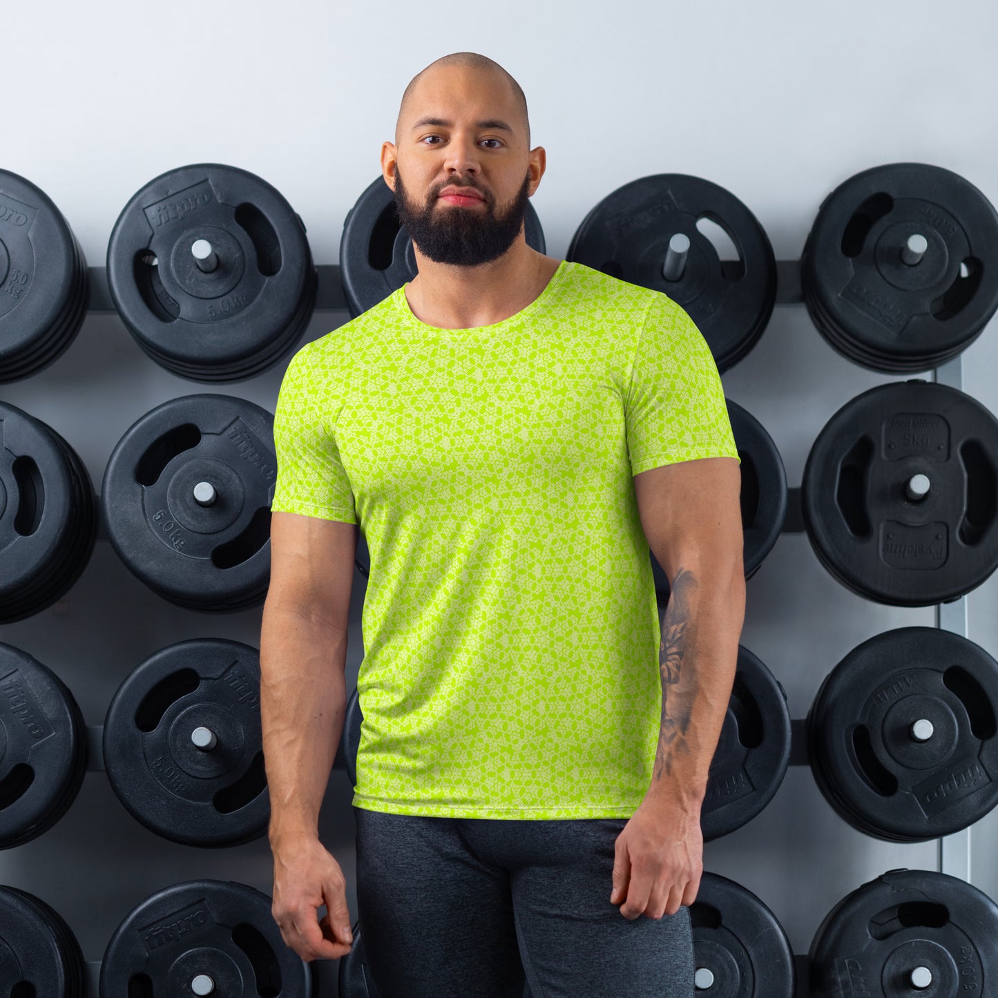 Athletic man standing in front of a gym weight plate rack wearing the t-shirt with the 'Heptagon' yellow-green fractal geometric pattern.