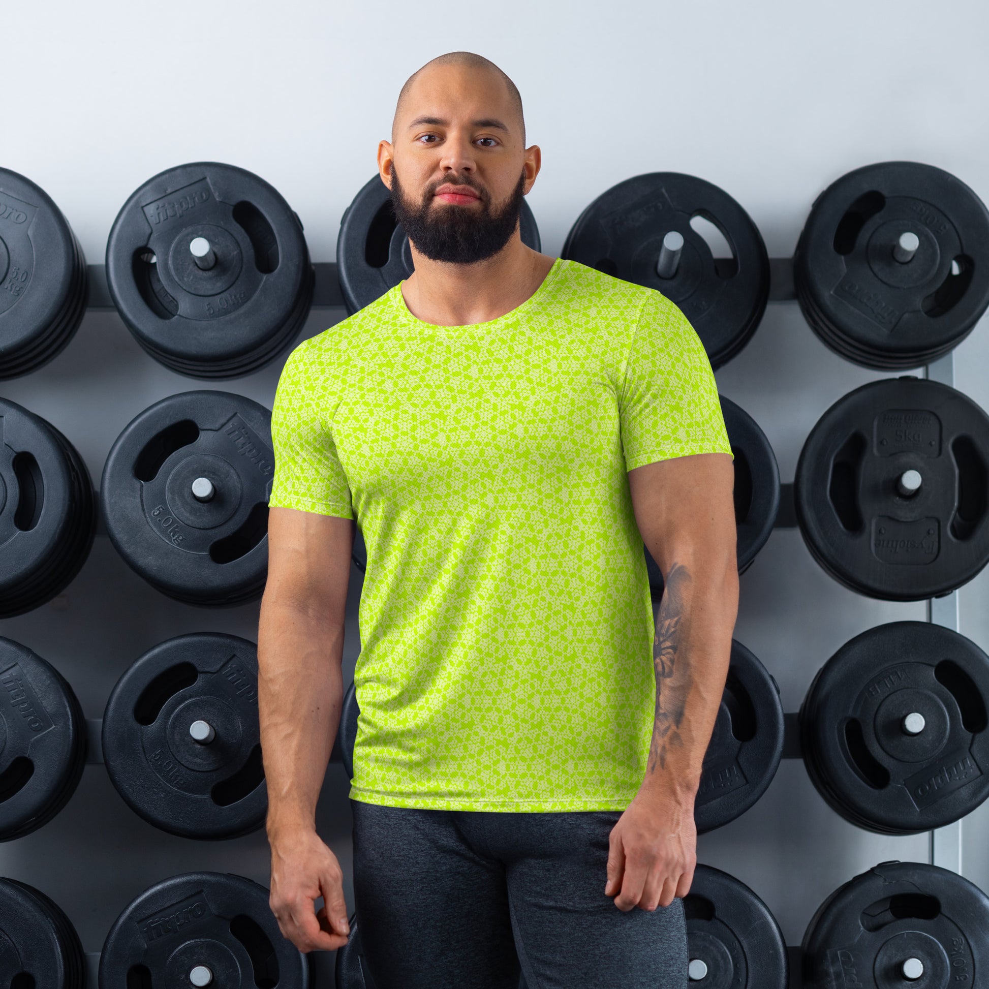 Athletic man standing in front of a gym weight plate rack wearing the t-shirt with the 'Heptagon' yellow-green fractal geometric pattern.