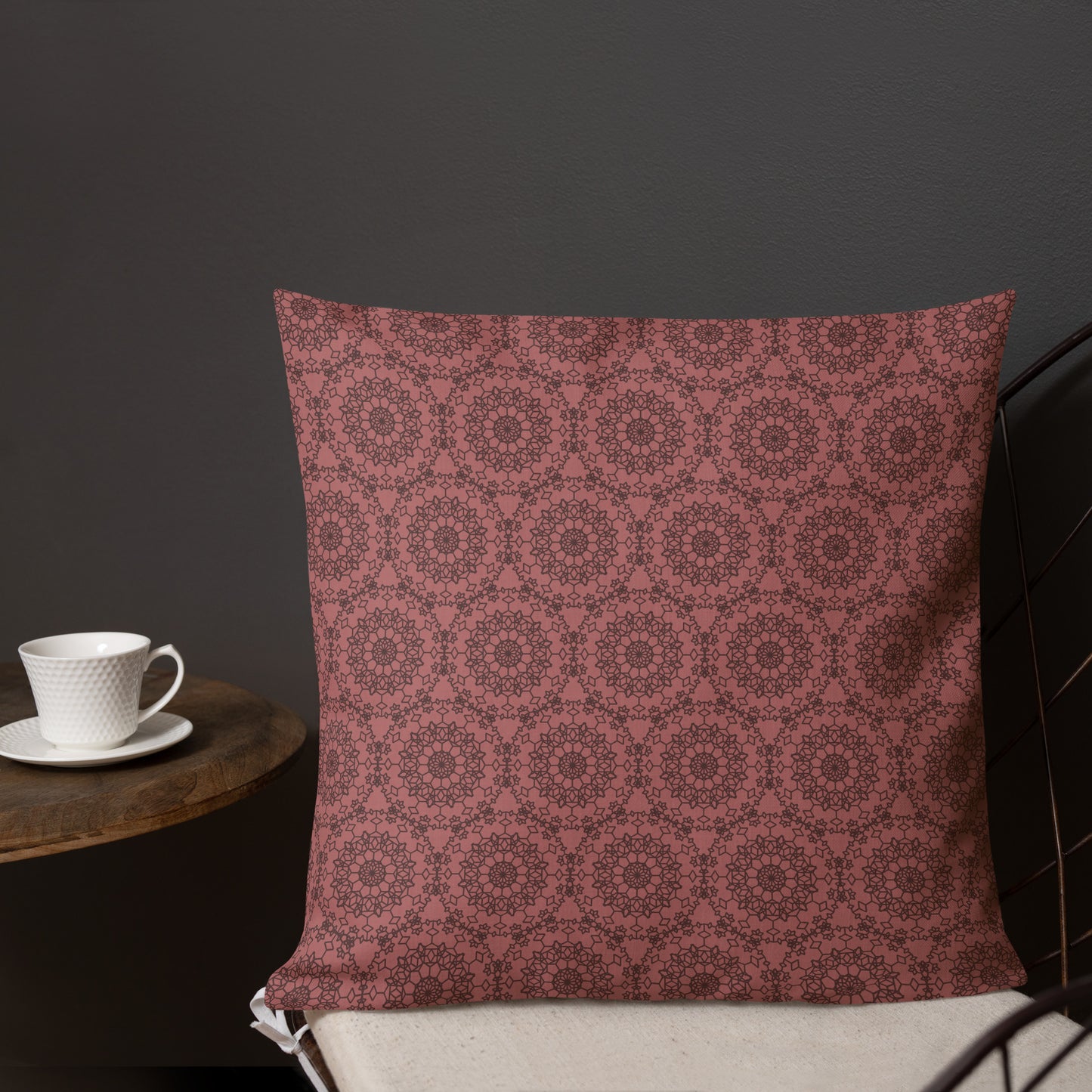 18″×18″ premium throw pillow with the 'Dodecagon' rose geometric pattern on wire chair beside side table with coffee cup.
