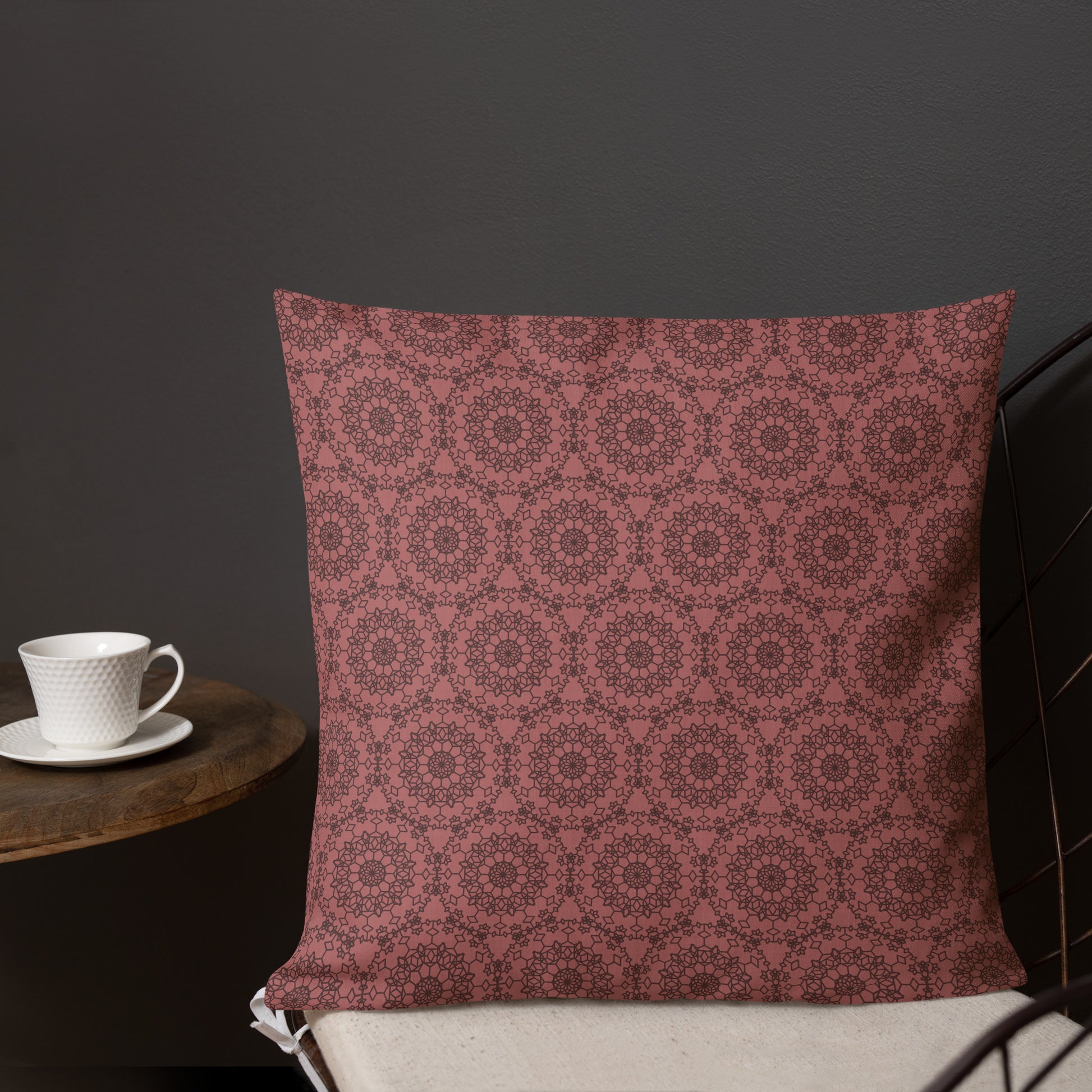 18″×18″ premium throw pillow with the 'Dodecagon' rose geometric pattern on wire chair beside side table with coffee cup.