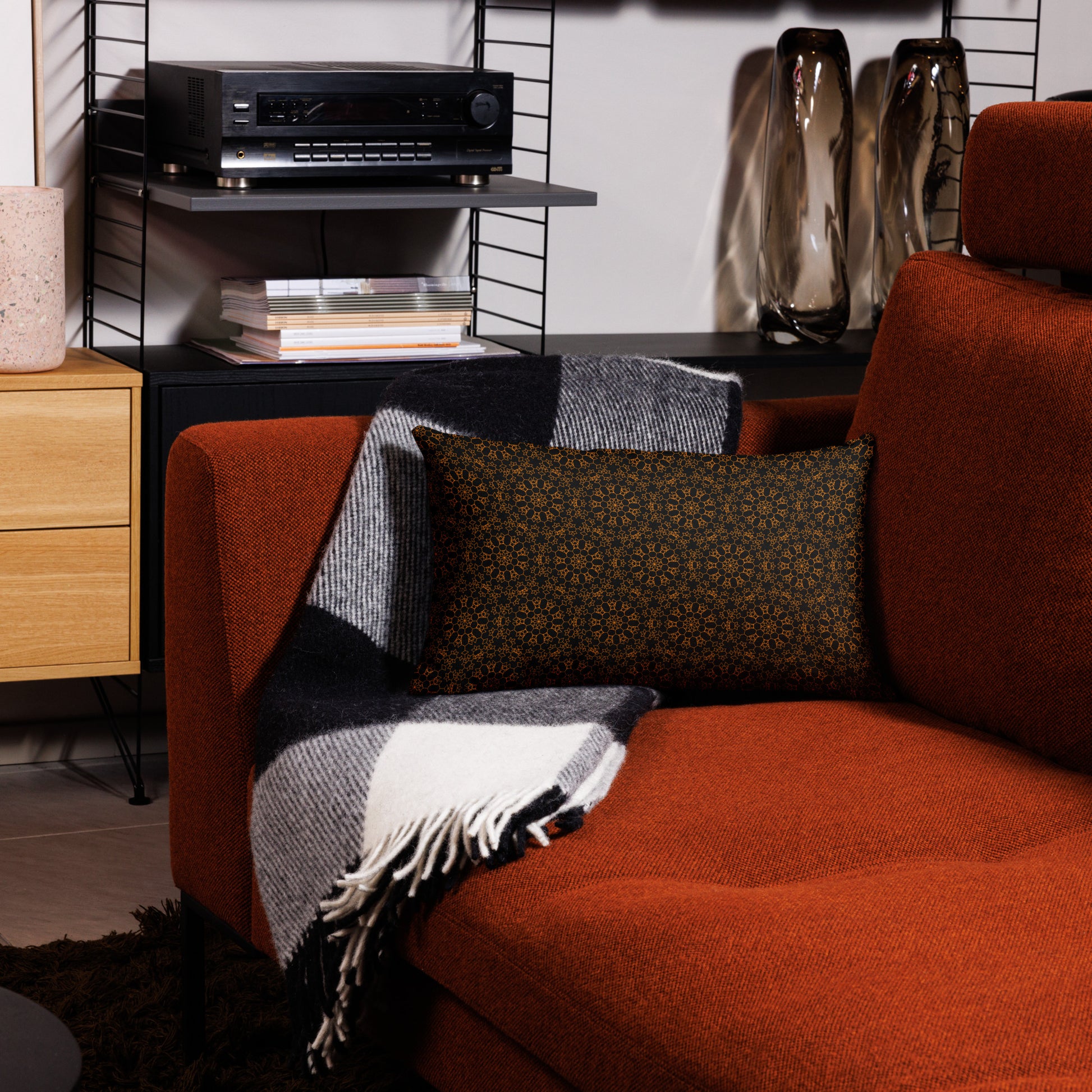20″×12″ premium throw pillow with the 'Enneagon' black & gold geometric pattern on a red armchair with checkered blanket.