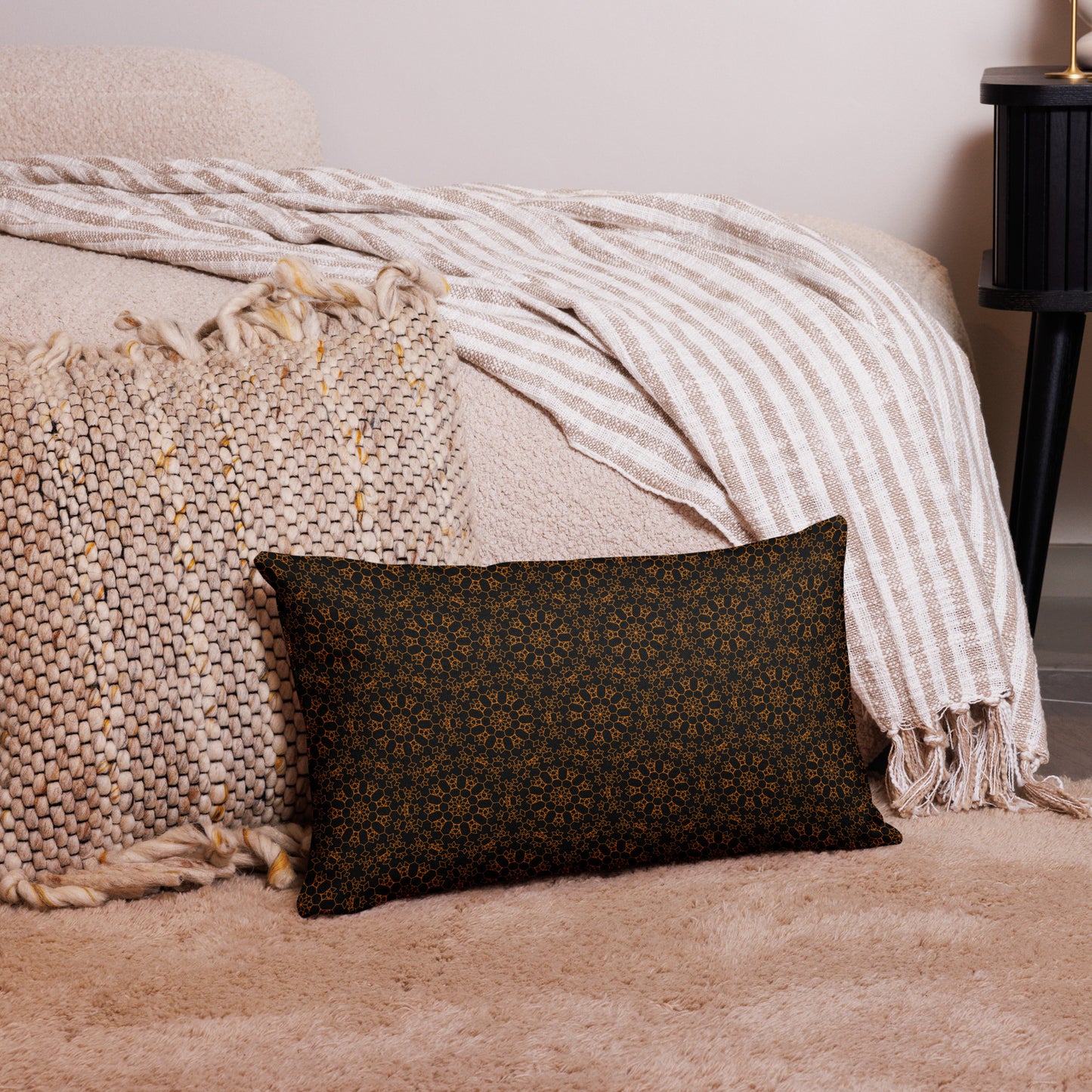20″×12″ premium throw pillow with the 'Enneagon' black & gold geometric pattern on a bed with striped blanket.