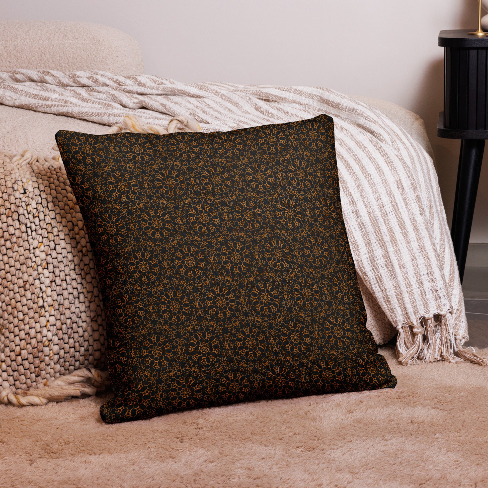 22″×22″ premium throw pillow with the 'Enneagon' black & gold geometric pattern on a bed with striped blanket.