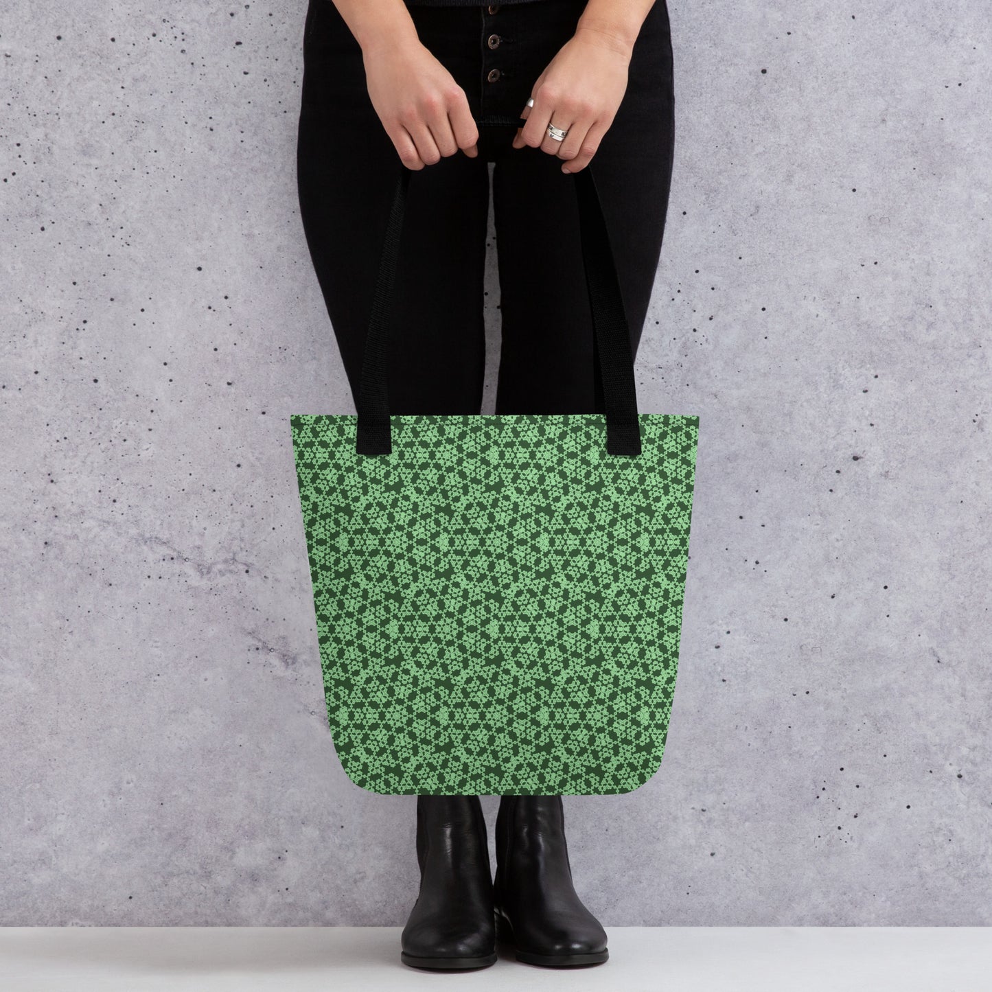 Close up of the lower part of a model holding with two hands the tote bag with the 'Heptagon' green fractal geometric pattern.