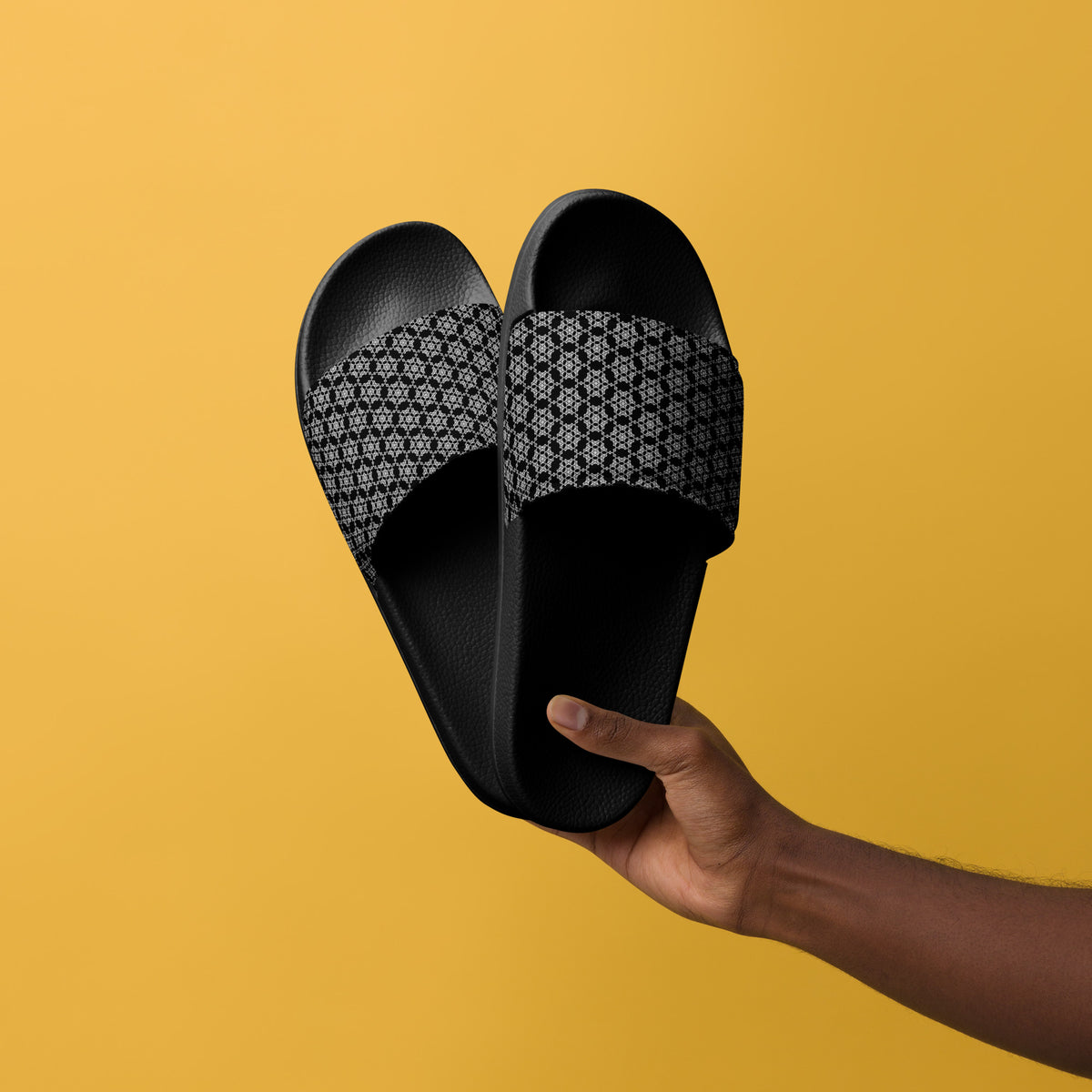 Hand holding a pair of the men's slides with the 'Hexagon' black and white fractal geometric pattern, against a yellow background.
