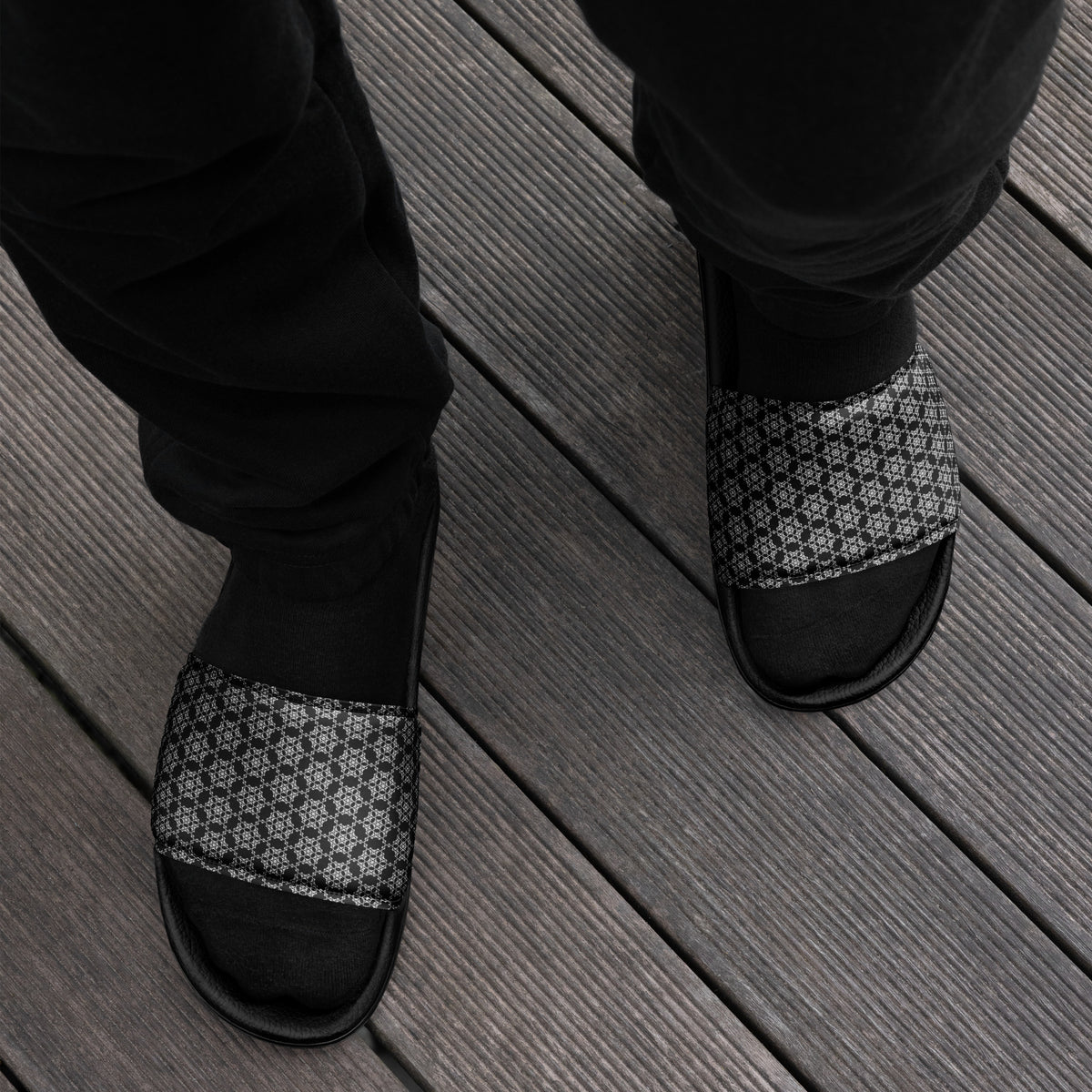 Top-down view of feet wearing the 'Hexagon' black and white fractal geometric pattern men's slides with black socks and black trousers on gray wooden decking.