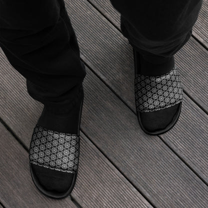 Top-down view of feet wearing the 'Hexagon' black and white fractal geometric pattern men's slides with black socks and black trousers on gray wooden decking.