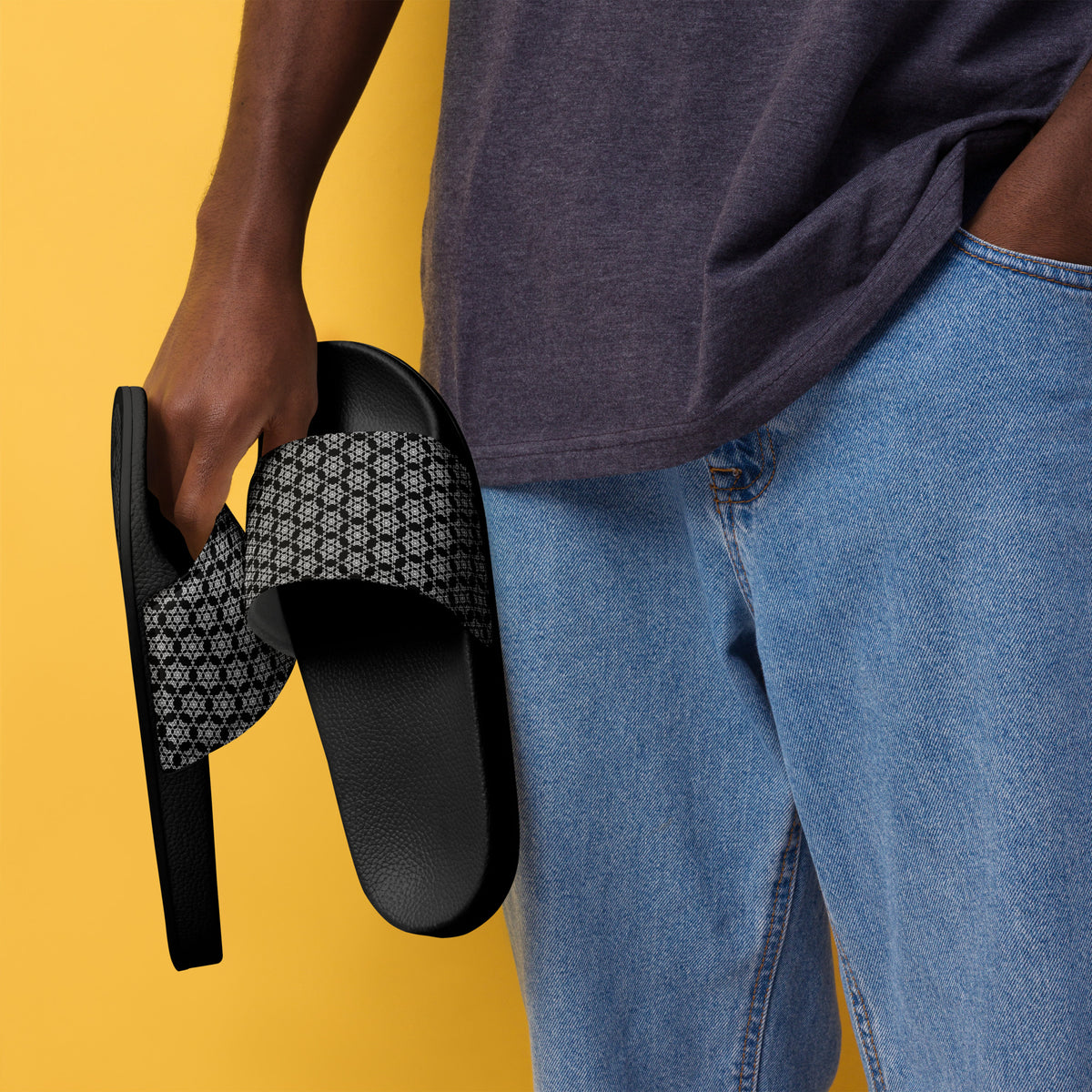 Man holding a pair of the men's slides with the 'Hexagon' black and white fractal geometric pattern, wearing a gray t-shirt and blue jeans, against a yellow background.