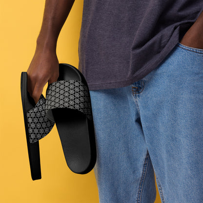 Man holding a pair of the men's slides with the 'Hexagon' black and white fractal geometric pattern, wearing a gray t-shirt and blue jeans, against a yellow background.